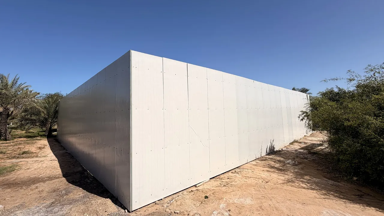 Exterior of a white insulated-panel facility shell on a sandy site, framed by palm trees and shrubs under a clear blue sky